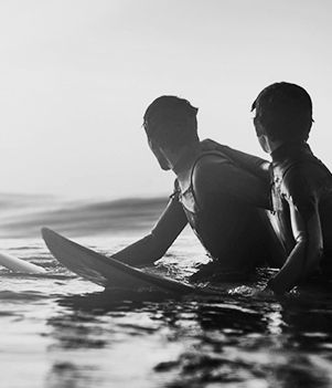 a black and white photo of a boy and man sitting on their surfboards looking to their right